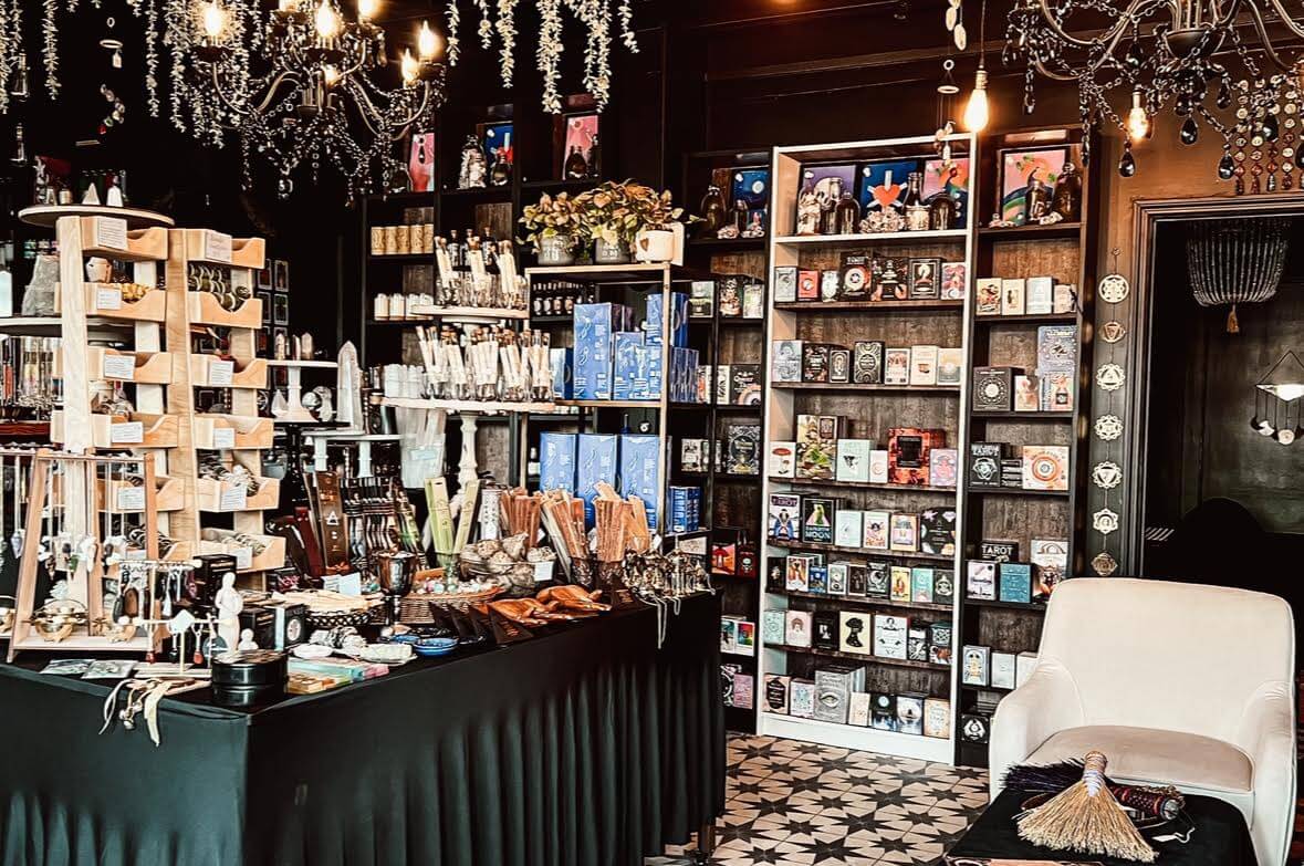 A boutique shop interior with shelves of tarot cards, books, jewelry, crystals, and decorative items; a white armchair and chandeliers are also visible—one of the best gift stores in Birmingham for unique finds.