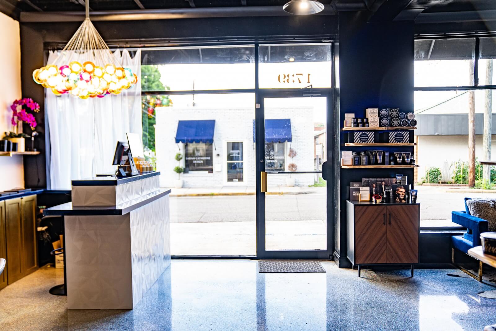 Welcome to one of the best hair salons in Birmingham, where our modern interior features a sleek reception desk, an elegant decorative light fixture, product shelves, and a glass door leading outside to relaxation. Indulge in premier spa services within this stylish setting.
