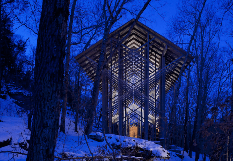 The glass and wood chapel, reminiscent of the renowned glass chapels of Arkansas, is beautifully illuminated at night. It stands serenely amidst snow-covered trees in a tranquil forest setting.