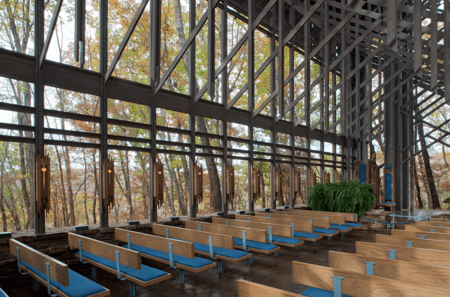 The interior of this chapel, reminiscent of the glass chapels of Arkansas, features wooden pews and large glass windows offering a breathtaking view of autumn trees outside.