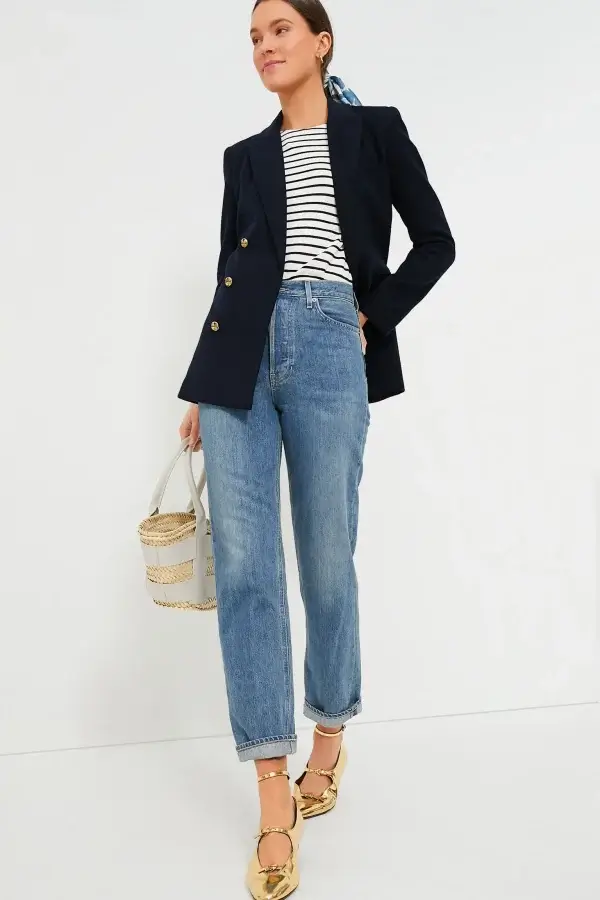 A person poses in a navy blazer and striped shirt—classic pieces given a modern spin with blue jeans, gold shoes, and a woven handbag—creating an ensemble perfect for spring wardrobe essentials.