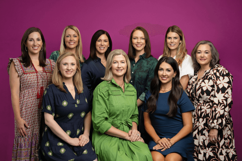 A group of nine women, featured in client-provided images, poses together against a purple backdrop at the Hutton Hotel, with five standing and four seated.