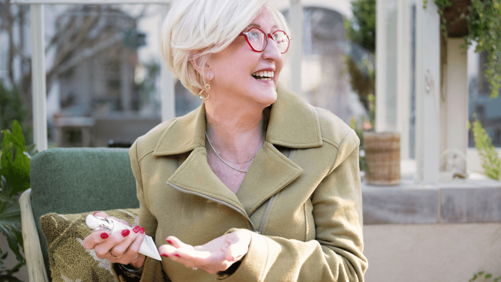 An older woman with short blonde hair and red glasses, wearing a green coat, smiles while holding a tissue. She is seated with plants in the background, embodying the warmth of Hutton Hotel in November 2024.