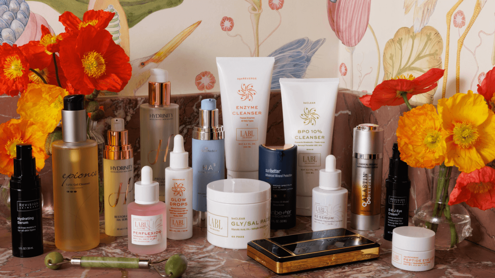 A collection of skincare essentials—cleansers, serums, and creams—gracefully arranged on a marble surface. The backdrop features colorful floral wallpaper and vibrant orange flowers, capturing the essence of elegance in the curated client-provided images for November 2024.