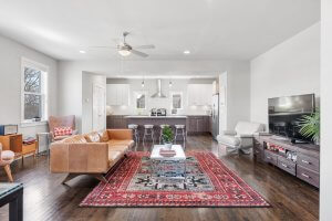 Discover an open living room on Nashville's charming 1517 Porter Rd, featuring a brown leather sofa and a patterned red rug beneath the wall-mounted TV. The kitchen, visible in the background, boasts white cabinets and stools. Enjoy the breeze from the ceiling fan and light streaming through large windows.