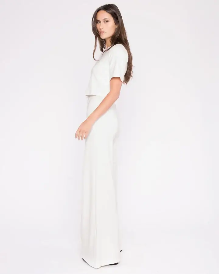A woman in a white two-piece outfit, a modern spin on classic pieces, stands against a plain white background. She poses sideways, looking towards the camera, with long hair draped over her shoulder. This ensemble is truly one of spring wardrobe essentials.
