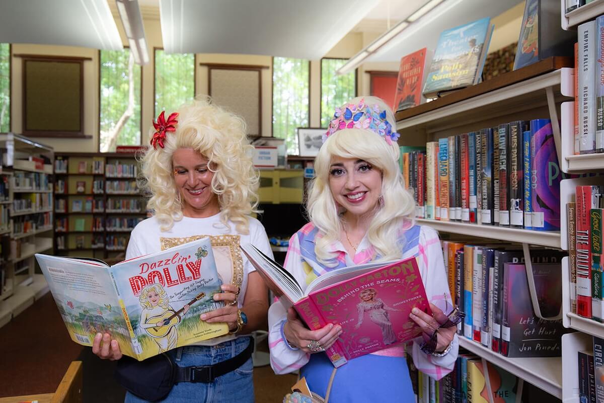 Two people wearing blonde wigs delve into a library's treasures, one engrossed in "Dazzlin' Dolly," an homage to the spirited Krewe of Dolly, while the other explores a book with a vibrant pink cover. Shelves brimming with stories stand proudly in the background.