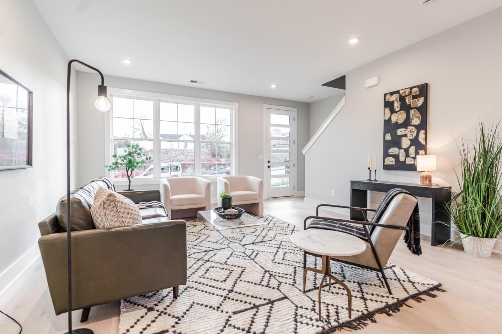 Modern living room on Trimble St, Nashville, with a large window, light gray walls, a patterned rug, a black sofa, two white chairs, a black side chair, dark desk with table lamp, tall plant, and floor lamp. A perfect blend of style and comfort in TN's vibrant heart.
