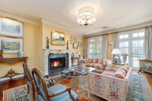 A cozy living room on West End Ave with a lit fireplace, floral sofas, and elegant chairs. Various framed paintings adorn the walls of this inviting Nashville space, featuring a chandelier and large windows draped with curtains.