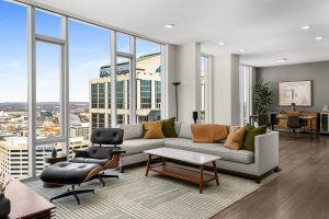 Discover a modern living room at 515 Church Street, Nashville, boasting large windows with city views, a gray sectional sofa, an Eames lounge chair, and a wooden coffee table. In the background, find an inviting dining area perfect for hosting.