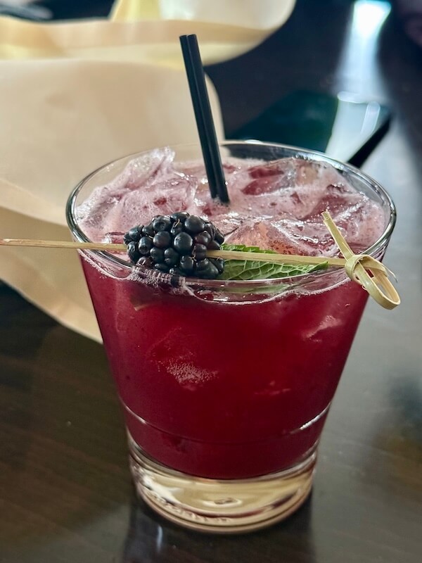 A refreshing glass of iced berry cocktail, infused with a hint of bourbon, garnished with a blackberry and mint, elegantly poised on a dark table with a straw.