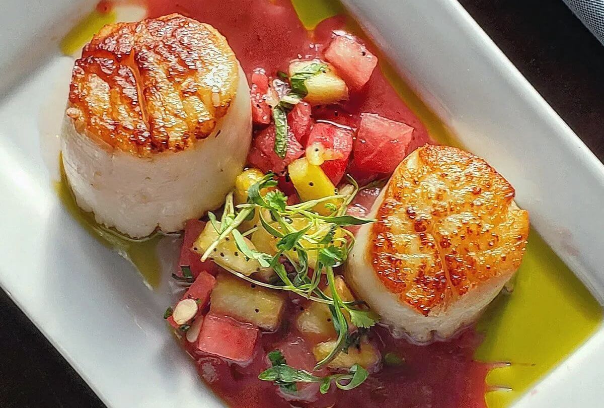 Two seared scallops on a sleek white plate, garnished with microgreens. They're nestled over a vibrant mix of diced watermelon and pineapple, elegantly finished with a drizzle of green sauce—perfect for Happy Hour indulgence.