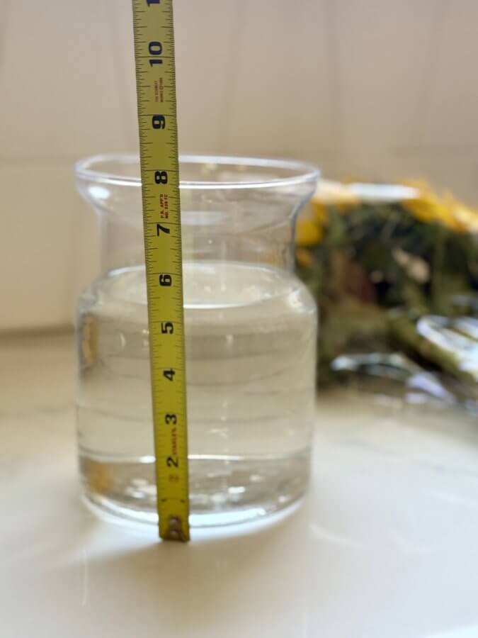 A measuring tape stands next to a clear water-filled glass jar, indicating a height of about 7 inches, while a floral arrangement of sunflowers brightens the blurred background.