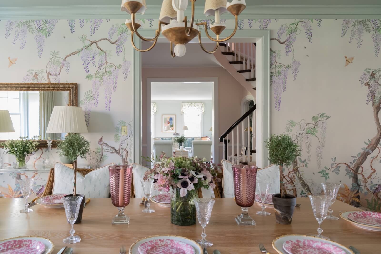 A dining room in tune with the best interior designers in Nashville, featuring floral wallpaper and a wooden table set with pink floral plates, crystal glassware, and pink vases. A floral centerpiece garnishes the table under a chandelier, while stairs ascend gracefully in the background.