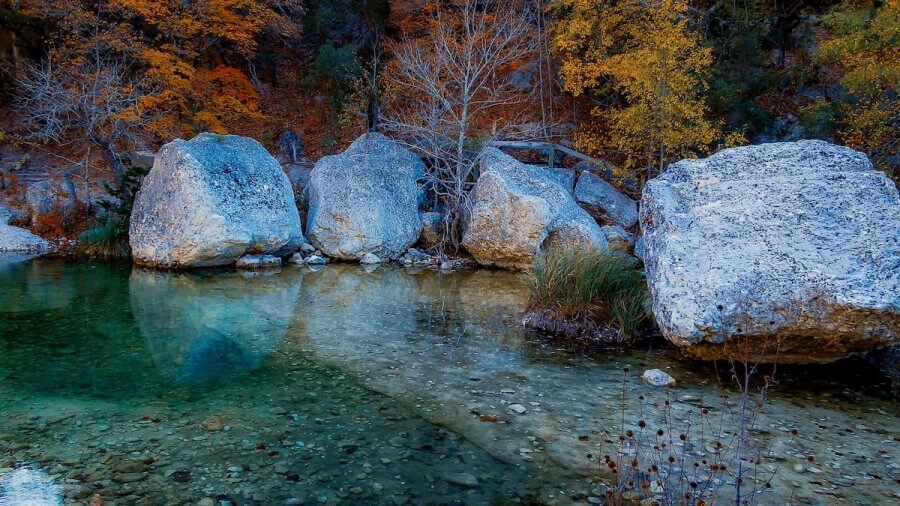 Clear water with visible stones flows by large boulders and autumn trees with orange and yellow leaves, reminiscent of serene Texas hikes.