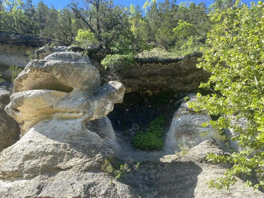 Explore this rocky landscape with large stone formations and an overhanging ledge, all surrounded by lush trees and greenery under a clear blue sky—a true gem for Texas hikes.