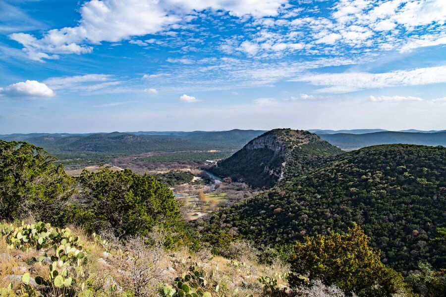 Discover the scenic view of a hilly landscape perfect for Texas hikes, where lush green vegetation meets a distant hill under a partly cloudy sky.