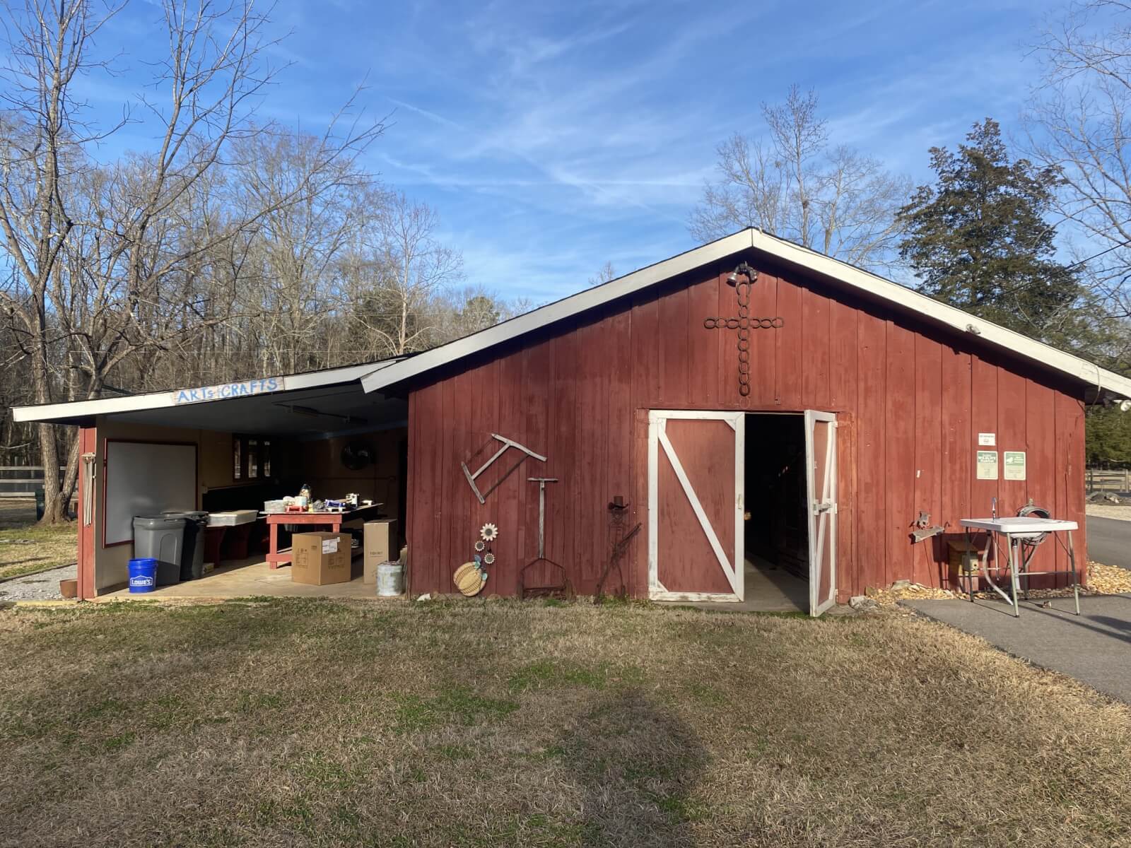 A red barn-like building with open doors reveals assorted items inside and out, evoking a sense of hope. Surrounded by trees under a clear blue sky.