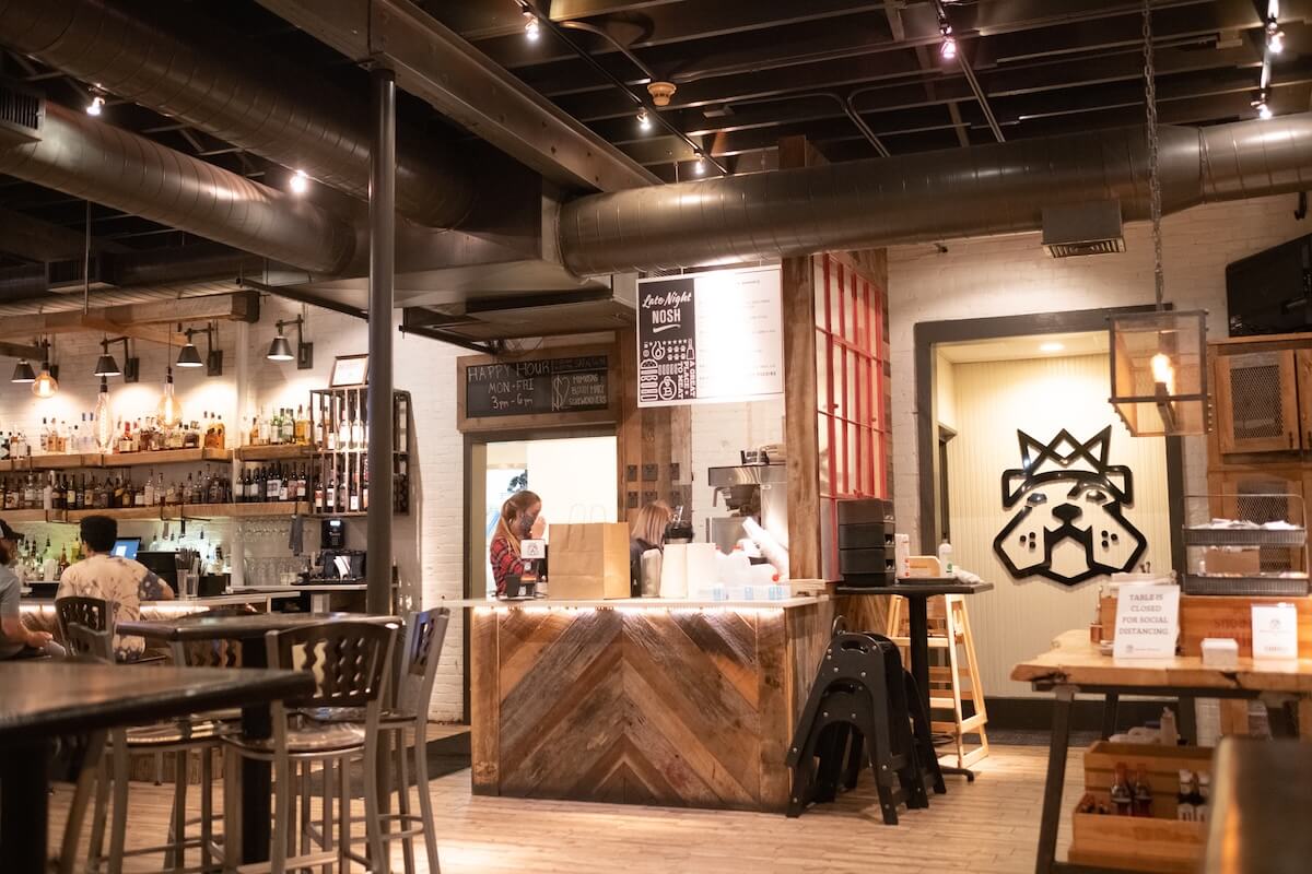 A modern, industrial-style bar in Starkville with two people behind the counter, bottles on shelves, warm lighting, and wooden accents. The space exudes a festive vibe with colorful art adorning the walls.
