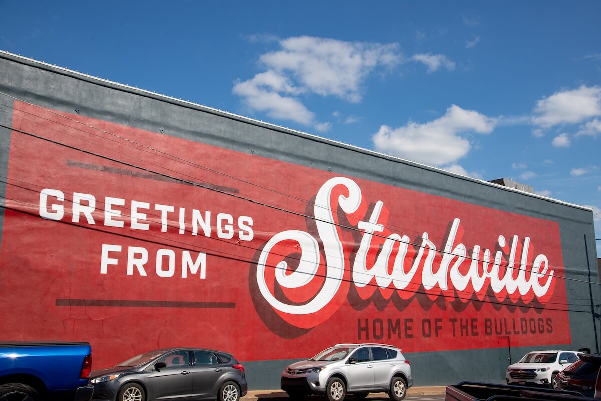 A large mural in Starkville, MS reads "Greetings from Starkville, Home of the Bulldogs." Cars are parked below this charming piece of art under a blue sky.