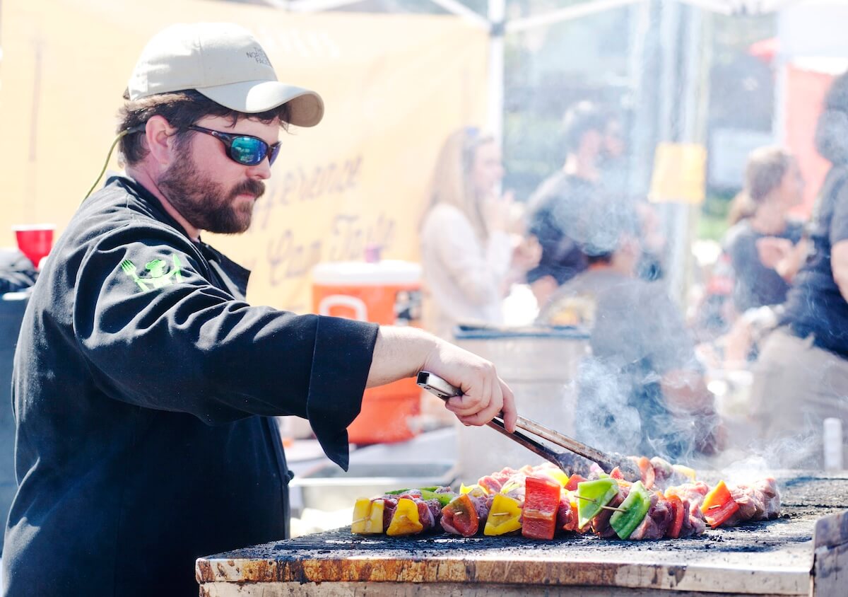At an outdoor event in Starkville, MS, a chef in a black jacket and cap expertly grills skewers of vegetables and meat amid lively crowds and bustling food stands.