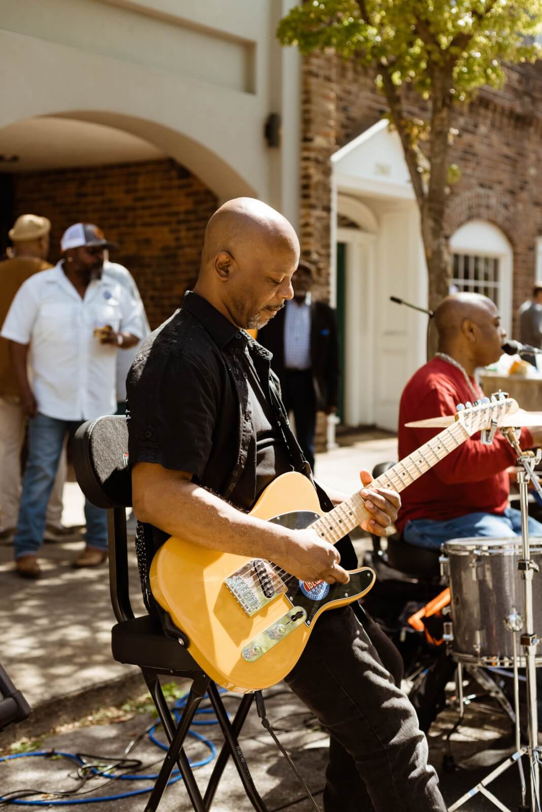 A musician strums an electric guitar on a stool outdoors, with another person playing drums nearby. Several people gather near a brick building, capturing the charm often found in small towns in Tennessee.
