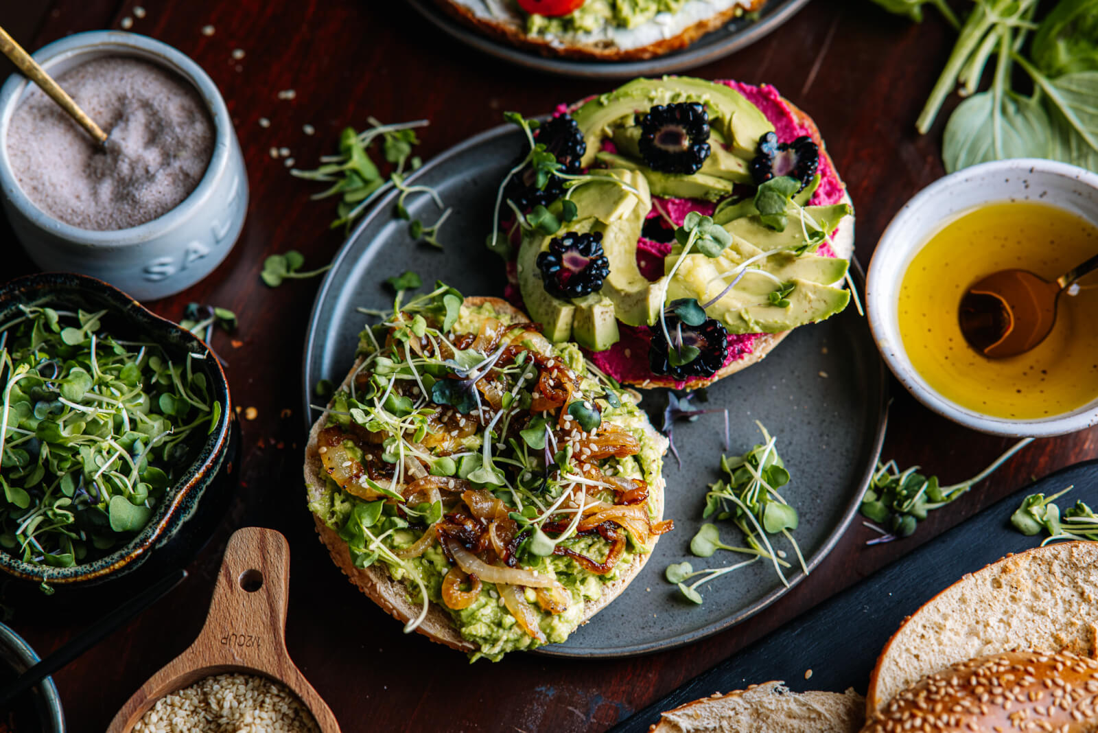 An avocado toast bar delight: two open-faced bagels adorned with creamy avocado, fresh sprouts, caramelized onions, berries, and greens on a plate. They’re paired perfectly with a bowl of oil, a dash of salt nearby, and an extra bowl of crisp sprouts for added crunch.