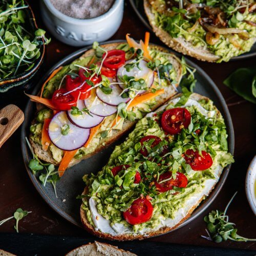 A plate of open-faced sandwiches serves as the centerpiece of an avocado toast bar, adorned with sliced radishes, cherry tomatoes, and microgreens. It's surrounded by bowls of salt, sesame seeds, and fresh greens on a dark wooden table.