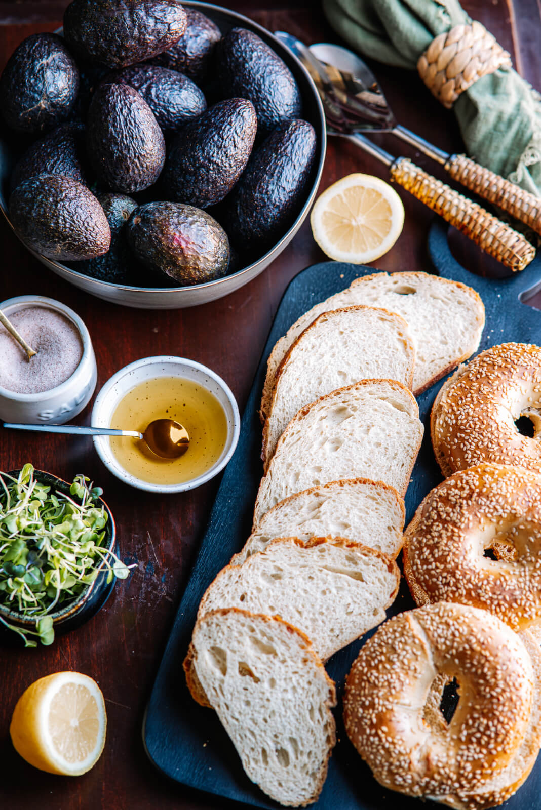 A breakfast spread transforms into an avocado toast bar, complete with sliced bread, sesame bagels, whole avocados, lemon halves, microgreens, olive oil, and a pepper bowl on a dark wooden table.