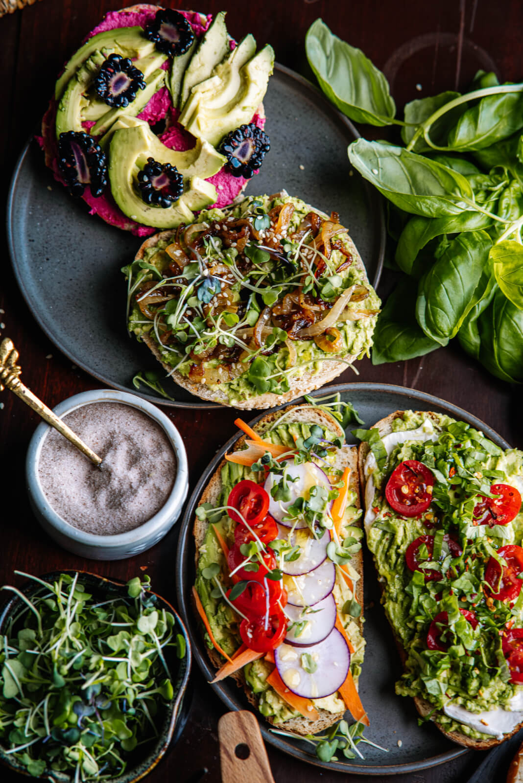 Assorted open sandwiches on an avocado toast bar feature toppings like tomatoes, radishes, microgreens, and caramelized onions. Fresh basil and salt are offered on the side.