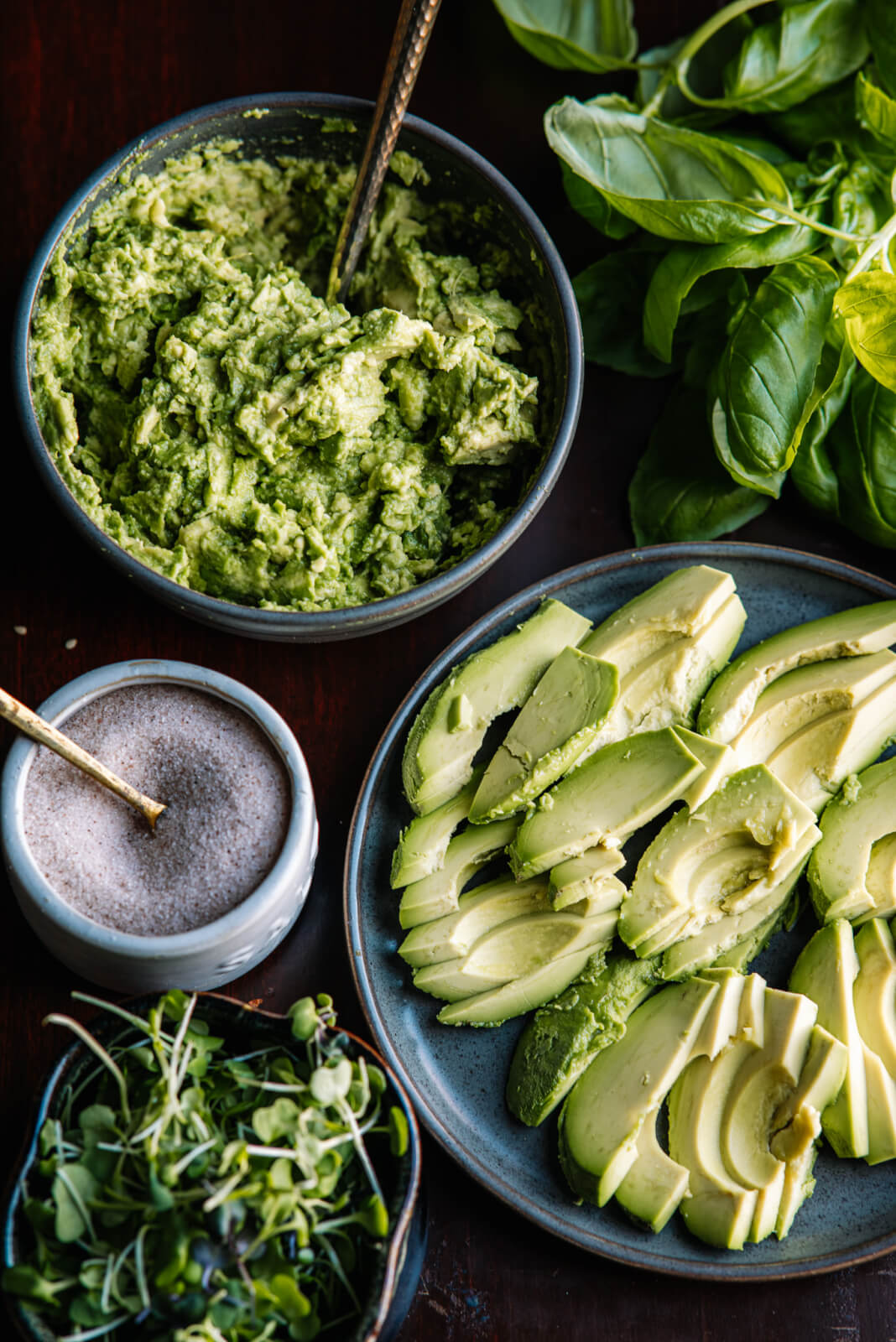 A delightful spread features a bowl of creamy avocado mash with a spoon, alongside sliced avocados on a plate, ready for your avocado toast bar. A small dish of salt, fresh basil leaves, and vibrant microgreens complete the scene on a dark surface.