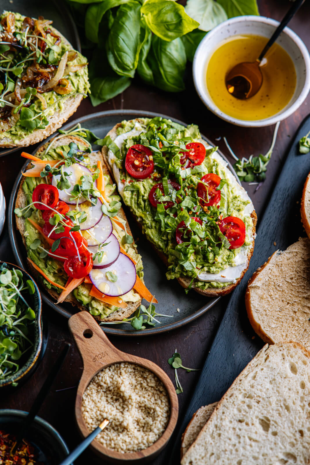 A delightful avocado toast bar featuring open-faced sandwiches with avocado, cherry tomatoes, radish slices, sprouts, and sesame seeds rests on a plate. Nearby, a bowl of olive oil and fresh greens adds to the inviting display.
