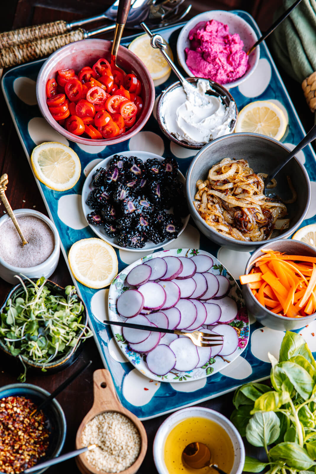 A vibrant avocado toast bar features an assortment of colorful ingredients on a tray, including sliced tomatoes, radishes, greens, lemon slices, cream cheese, beet hummus, sautéed onions, and shredded carrots, complemented by bowls of enticing spices.