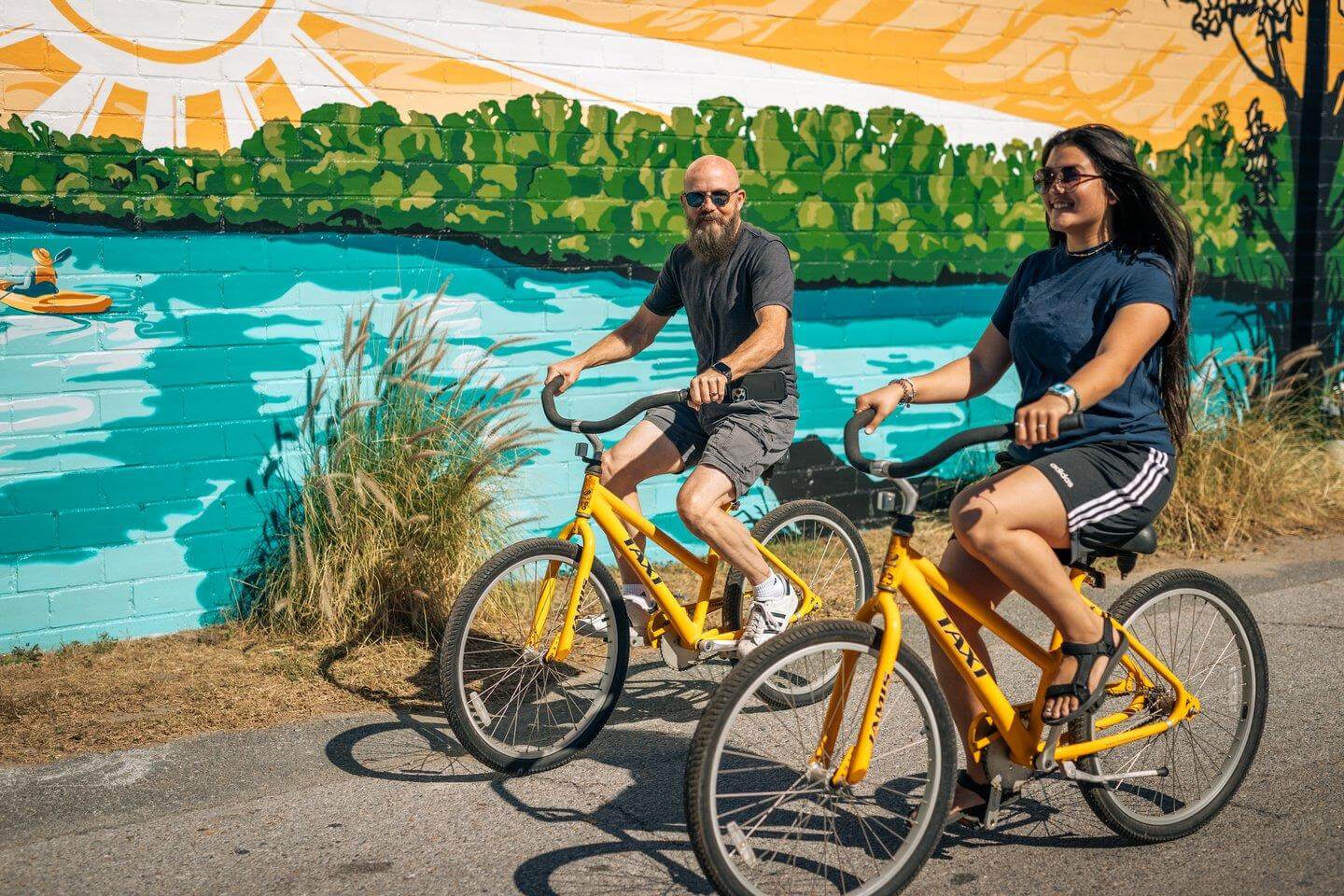 7 Best Family Vacations for Outdoor Fun - 18 Two people enjoy outdoor fun as they ride yellow bikes along a path adorned with a colorful mural of greenery and a sunlit sky.