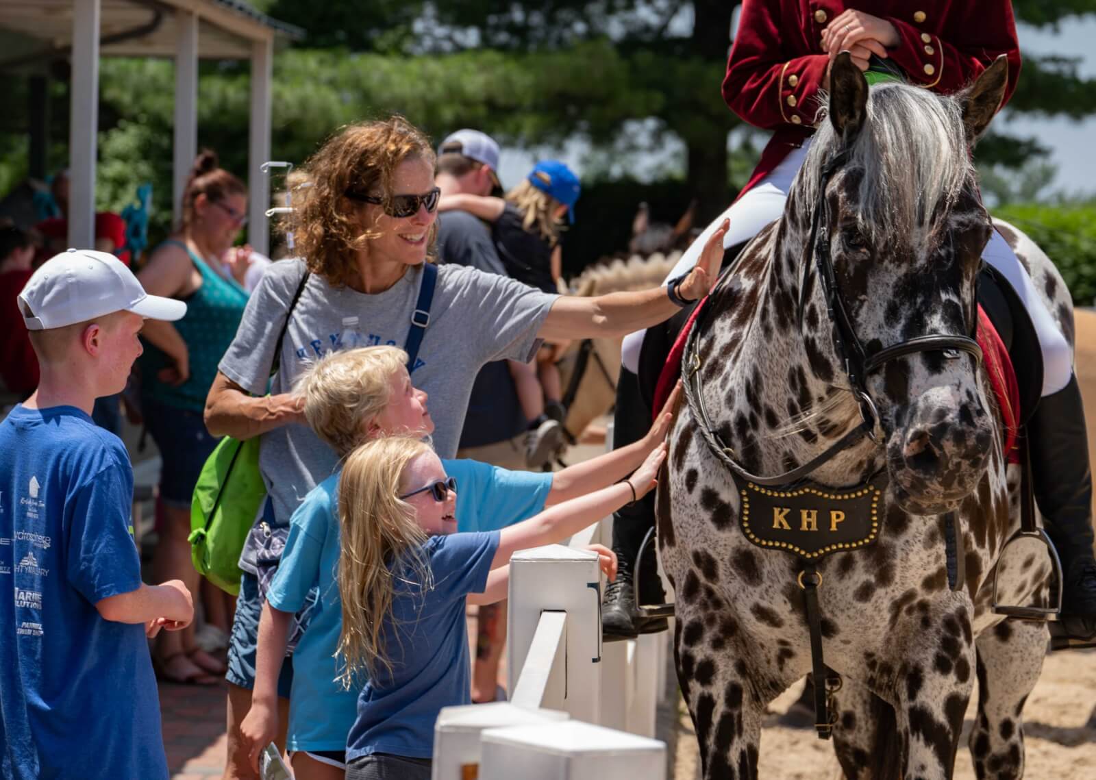7 Best Family Vacations for Outdoor Fun - 13 A woman and two children enjoy some outdoor fun as they pet a dappled horse with "KHP" on its bridle. A rider is seated on the horse, while people gather in the background under a shaded area.