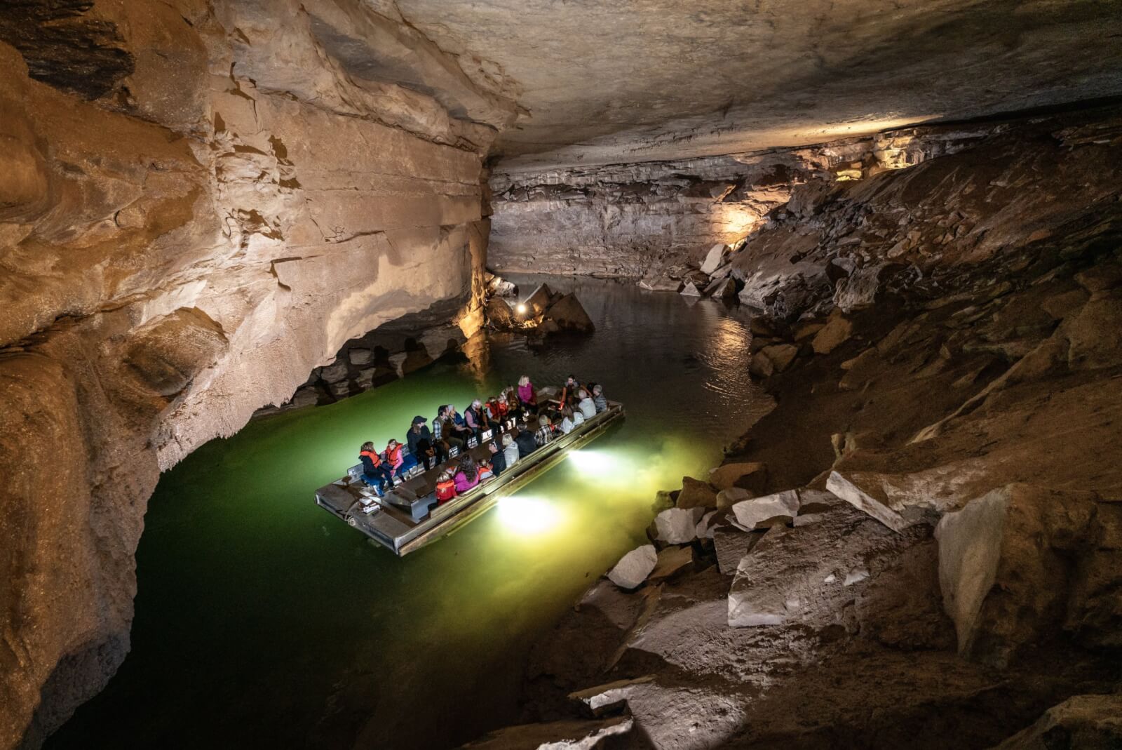 7 Best Family Vacations for Outdoor Fun - 8 A group of people enjoy outdoor fun in a boat, traveling through an underground cave with rocky walls and a greenish water surface, illuminated by artificial lights.