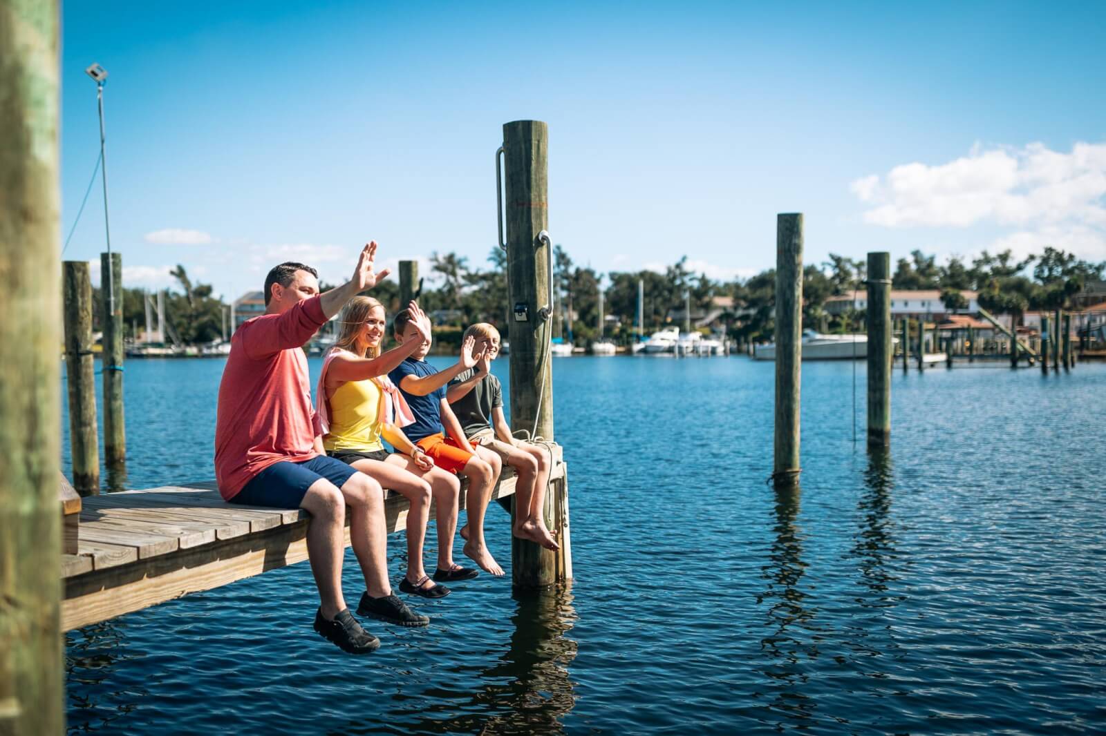 7 Best Family Vacations for Outdoor Fun - 17 Four people sit on a wooden dock overlooking the water, waving happily. It's a sunny day filled with family vacations vibes, as boats glide by and palm trees sway gently in the background, epitomizing outdoor fun.