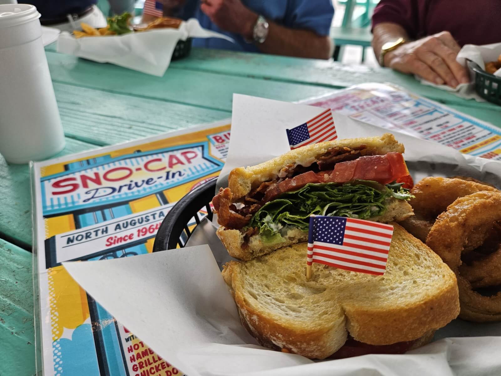 7 Best Family Vacations for Outdoor Fun - 20 Sandwich with lettuce, tomato, and bacon, topped with a small American flag toothpick, served in a basket with onion rings on a turquoise table. Perfect for outdoor fun during family vacations at the iconic Sno-Cap Drive-In, where memories mix with mouthwatering bites.