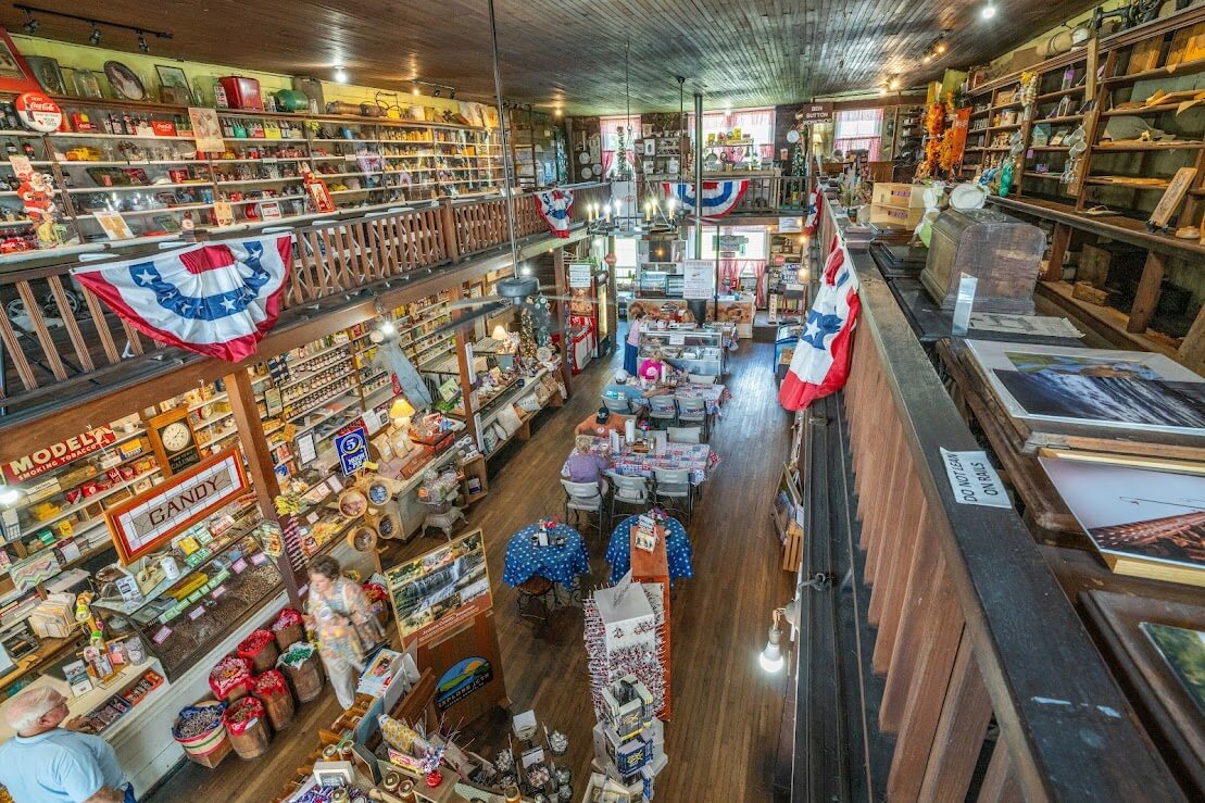 6 Southern Outdoor Getaways for the Adventure Traveler - 9 A rustic general store interior with wooden floors and shelves filled with assorted goods, perfect for the adventure traveler. Two levels with a balcony, red, white, and blue bunting, and various products evoke the charm of Southern outdoor getaways.