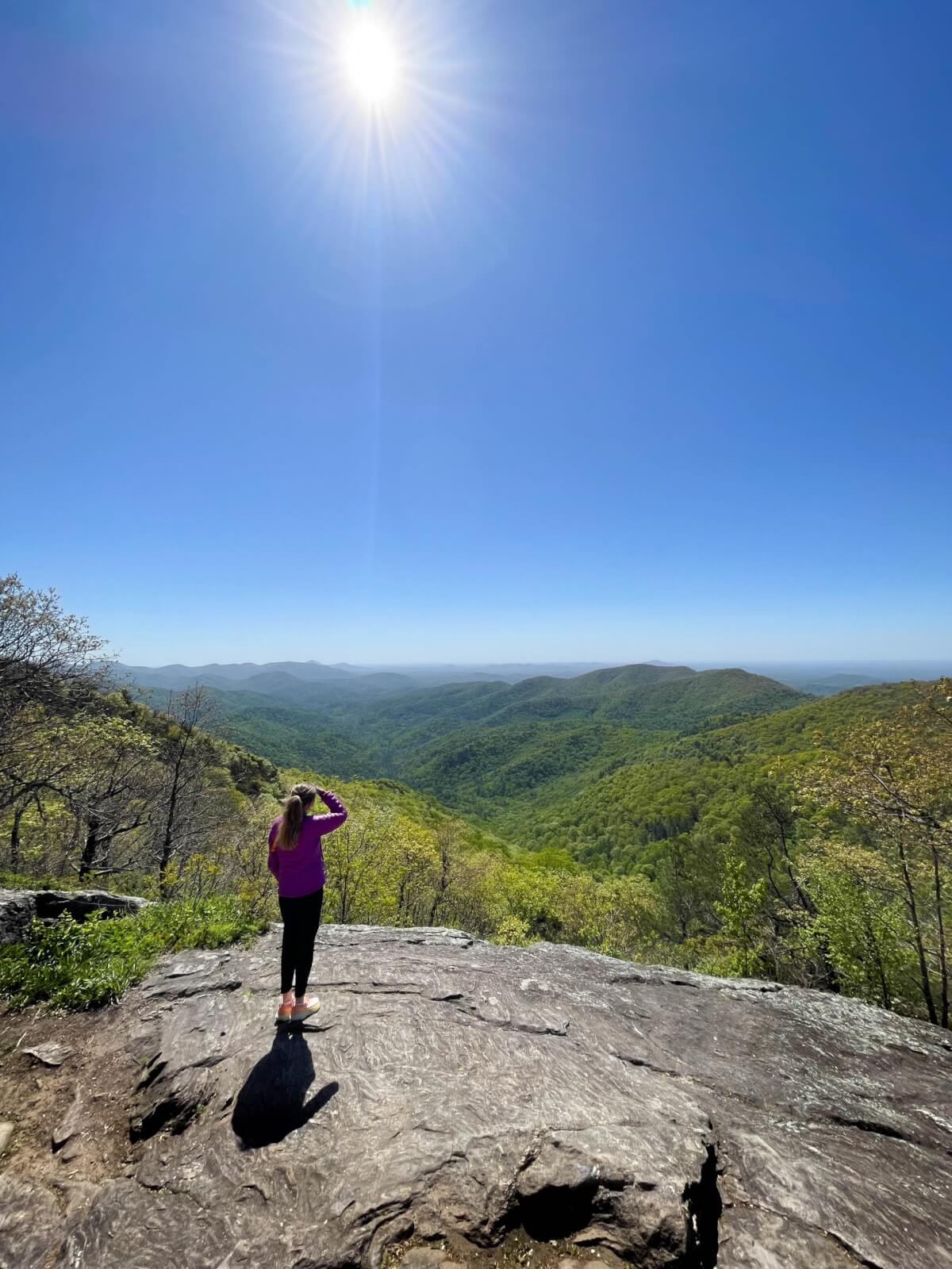 6 Southern Outdoor Getaways for the Adventure Traveler - 2 A person in a pink top stands on a rock, embodying the spirit of an adventure traveler, gazing at a vast green valley and distant hills under a bright sun and clear blue sky.
