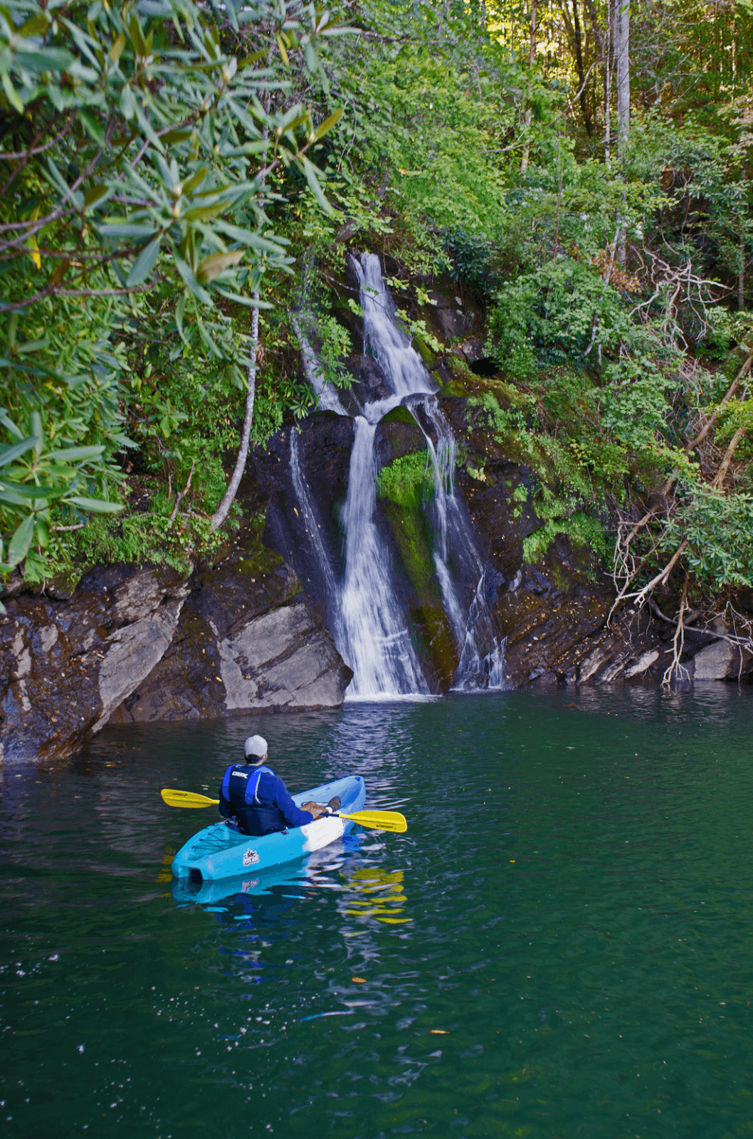 6 Southern Outdoor Getaways for the Adventure Traveler - 11 An adventure traveler in a blue kayak paddles on a forested lake towards a cascading waterfall, surrounded by lush greenery, capturing the essence of Southern outdoor getaways.