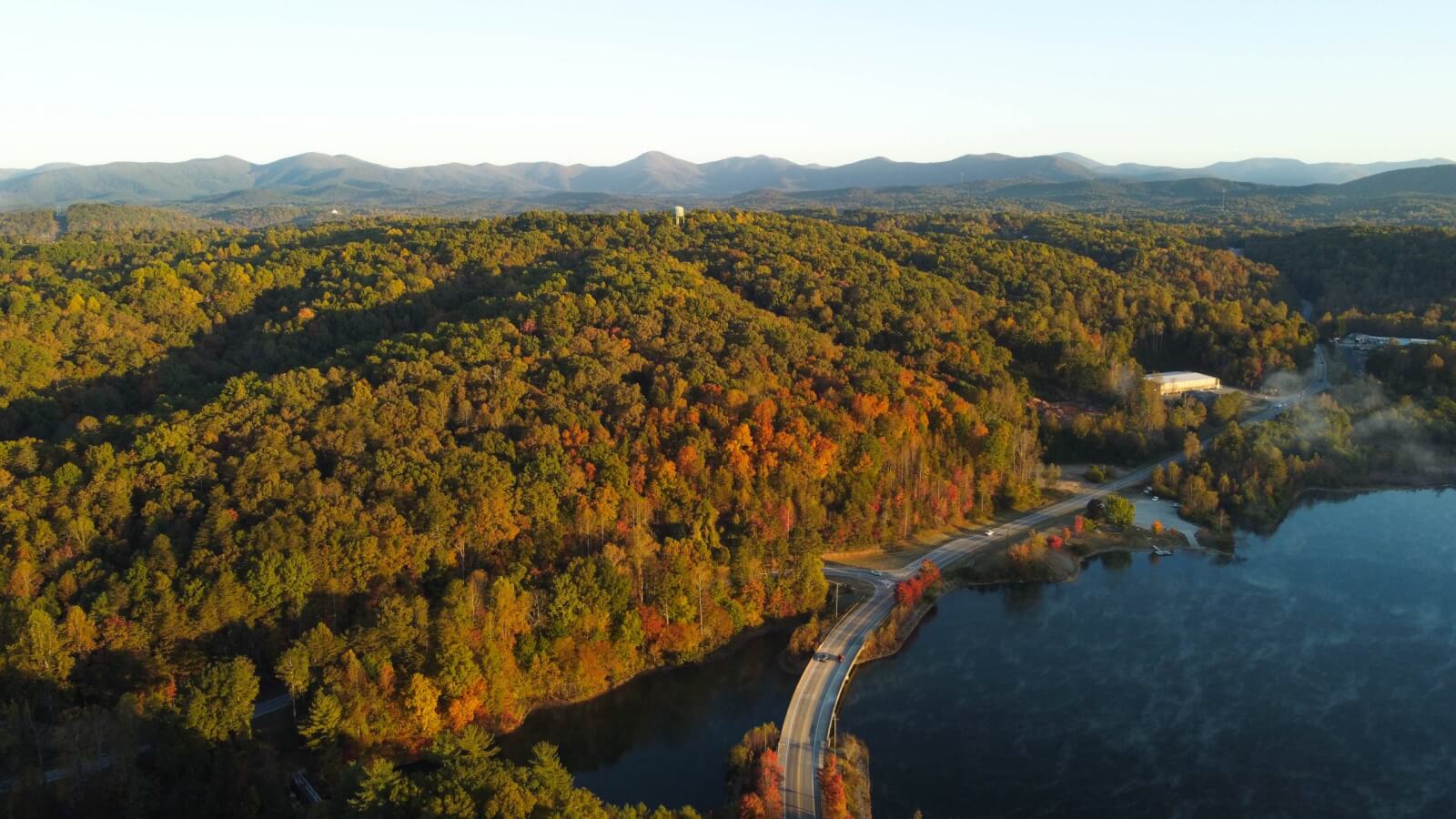 6 Southern Outdoor Getaways for the Adventure Traveler - 3 Aerial view of a Southern Outdoor Getaway: a forested hillside with autumn foliage, a winding road, and a lake in the foreground, set against distant mountains under a clear sky. Perfect for the adventure traveler seeking breathtaking vistas.
