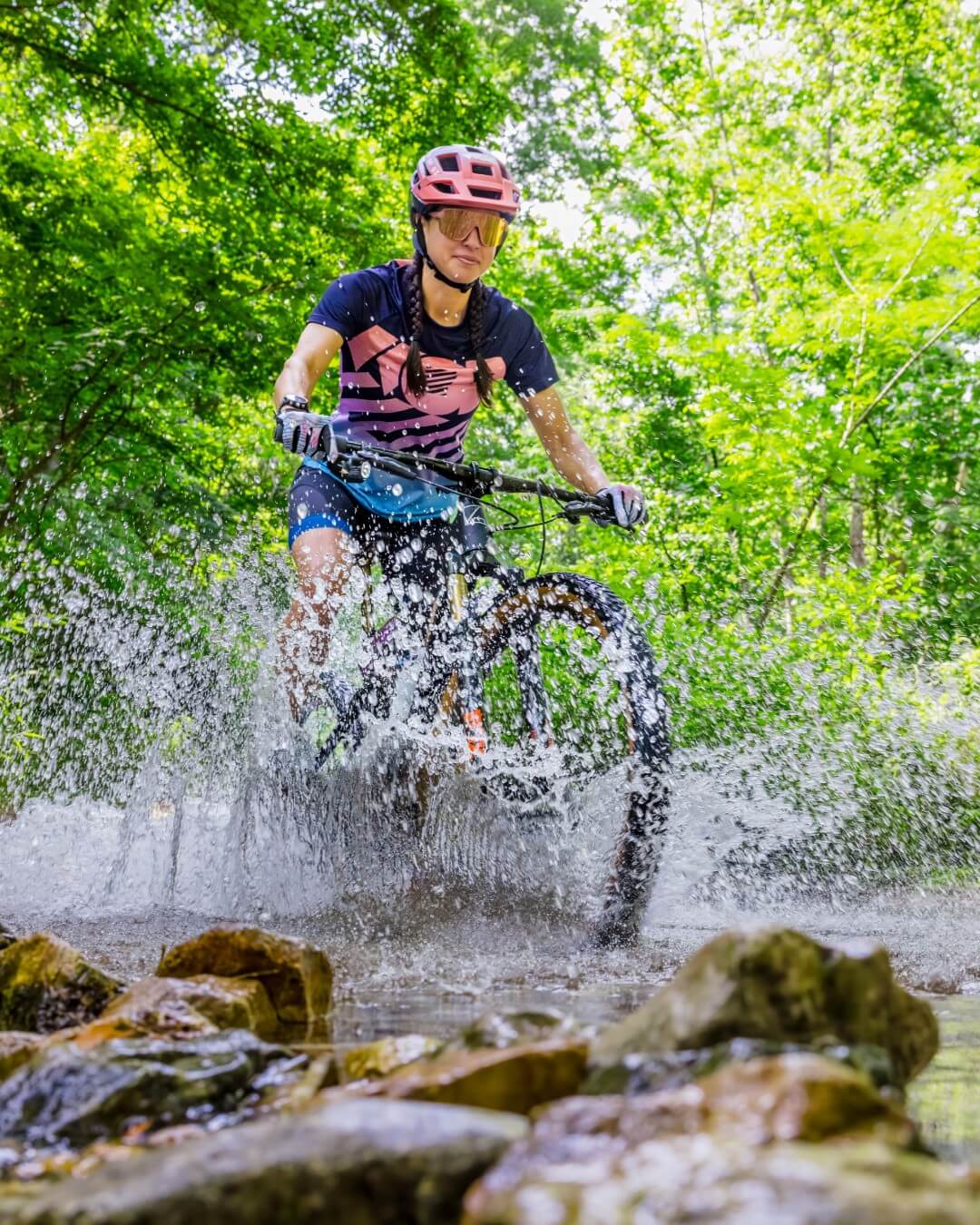 6 Southern Outdoor Getaways for the Adventure Traveler - 16 Cyclist in pink helmet and black outfit rides through a shallow stream, splashing water in a green, wooded area. This outdoor adventure embodies the thrill seekers of Southern Outdoor Getaways.