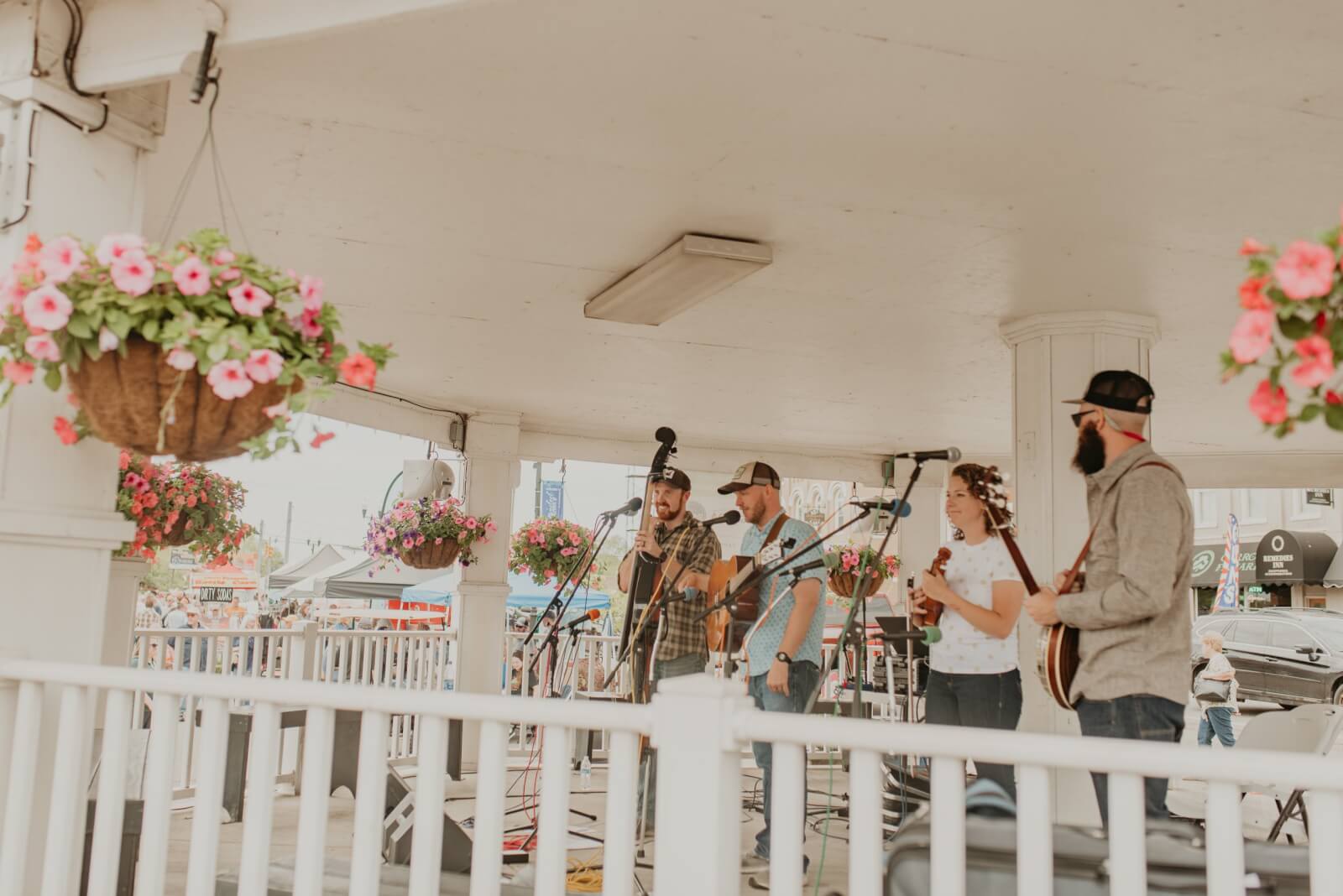 In a quaint Tennessee town, a band performs on a charming gazebo stage with flowers hanging nearby. Four musicians play various instruments, including a banjo and upright bass, while a small audience in the heart of the historic downtown enjoys the melodic tunes.