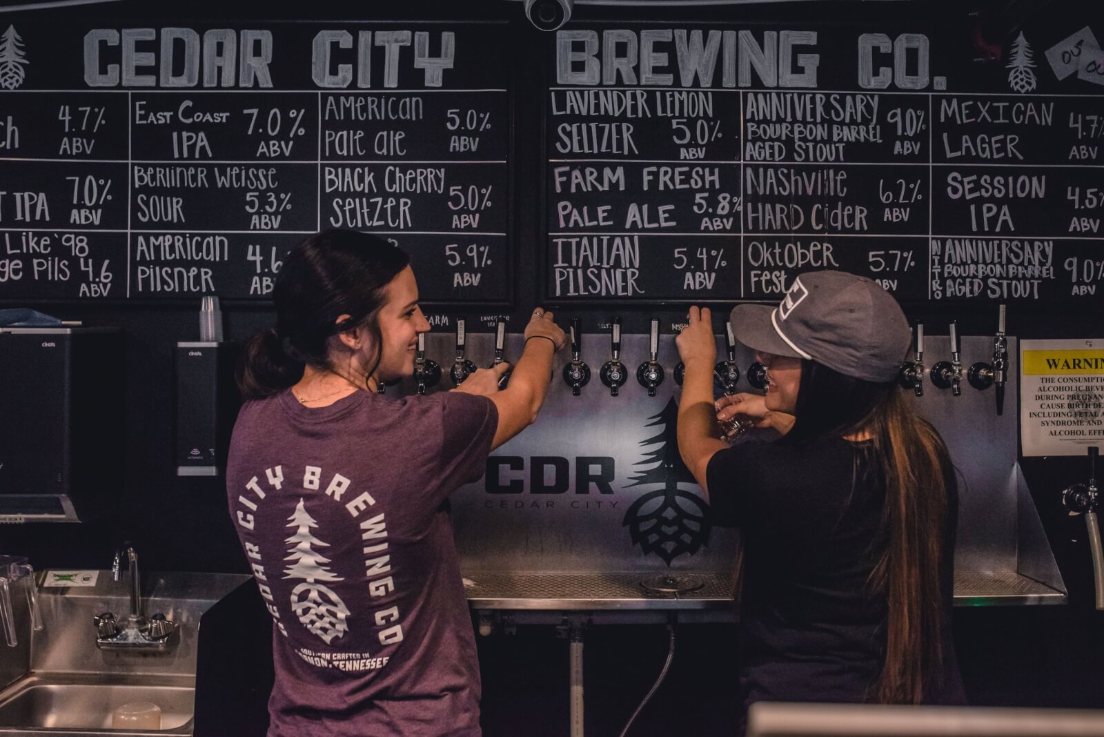 Two people pour drinks from taps at Cedar City Brewing Co. in the historic downtown. A menu above lists various beverages and their alcohol content.