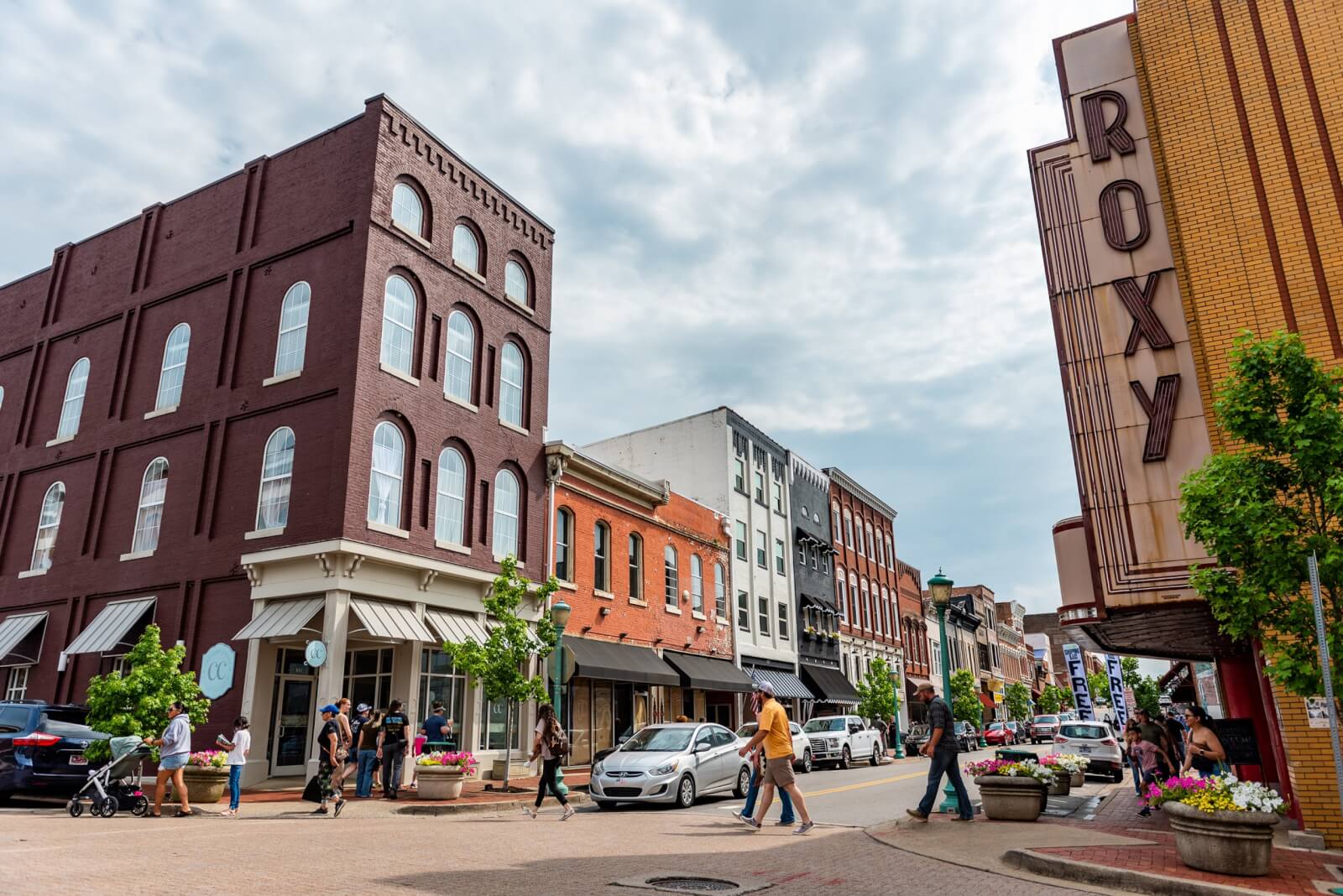 A lively street scene unfolds in downtown Clarksville, Tennessee, with people strolling past a row of historic buildings. Cars line the side, while a theater marquee boldly reads "ROXY" under a partly cloudy sky.