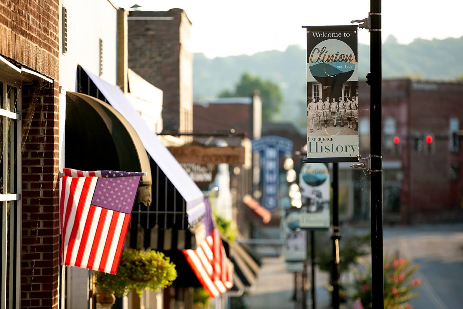 A vibrant street view of a small town showcases American flags, a "Welcome to Clinton" sign, and charming brick buildings of Tennessee's historic downtowns. Bright sunlight floods the scene, highlighting its timeless charm.