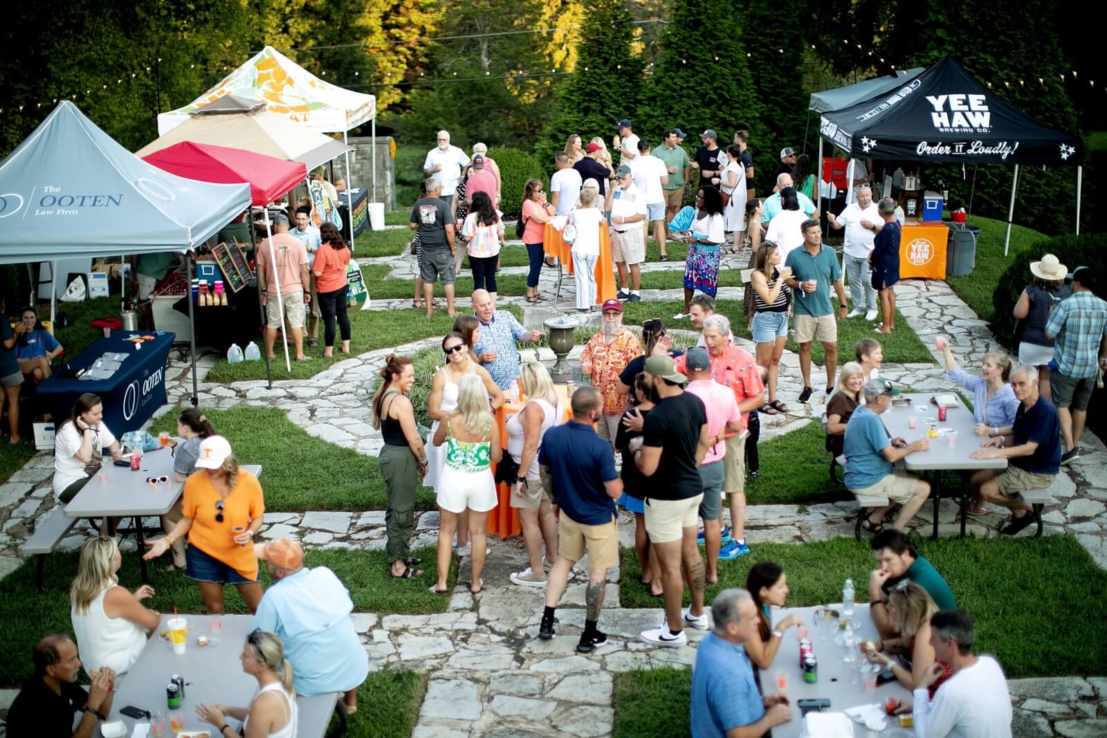 A community gathering outdoors with people socializing near tents in a historic Tennessee setting. Tables with chairs are set up on a grassy area intersected by a stone pathway.
