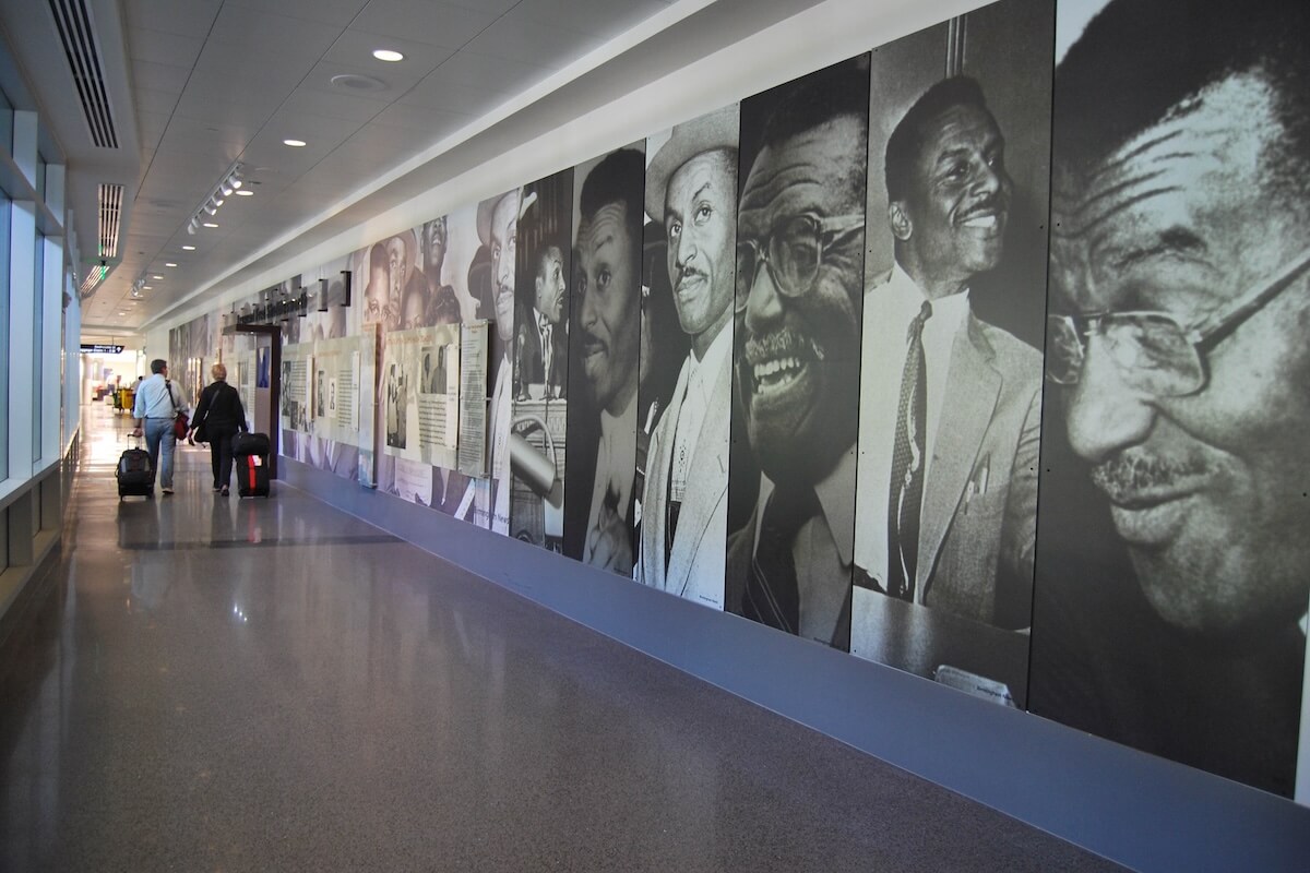 People walk through an airport corridor lined with large black-and-white portraits and informational panels about prominent individuals, perhaps exploring while waiting for their direct flights from Birmingham.