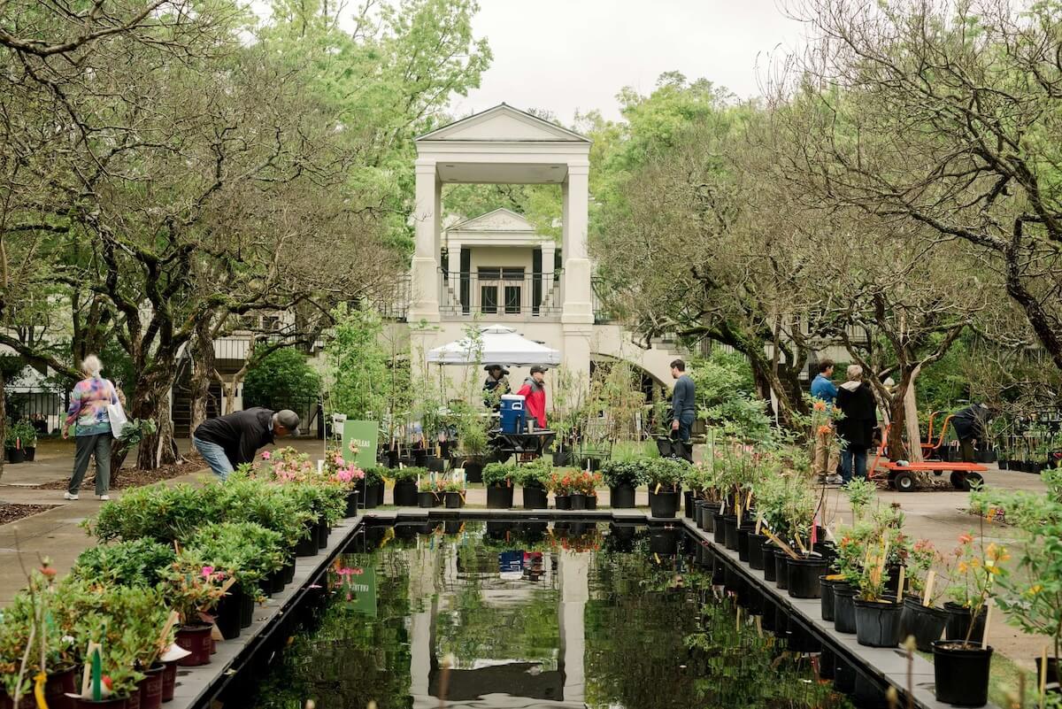 People browse potted plants arranged around a rectangular reflecting pool in an outdoor garden setting at a Birmingham events April showcase, with a white pavilion and trees in the background.